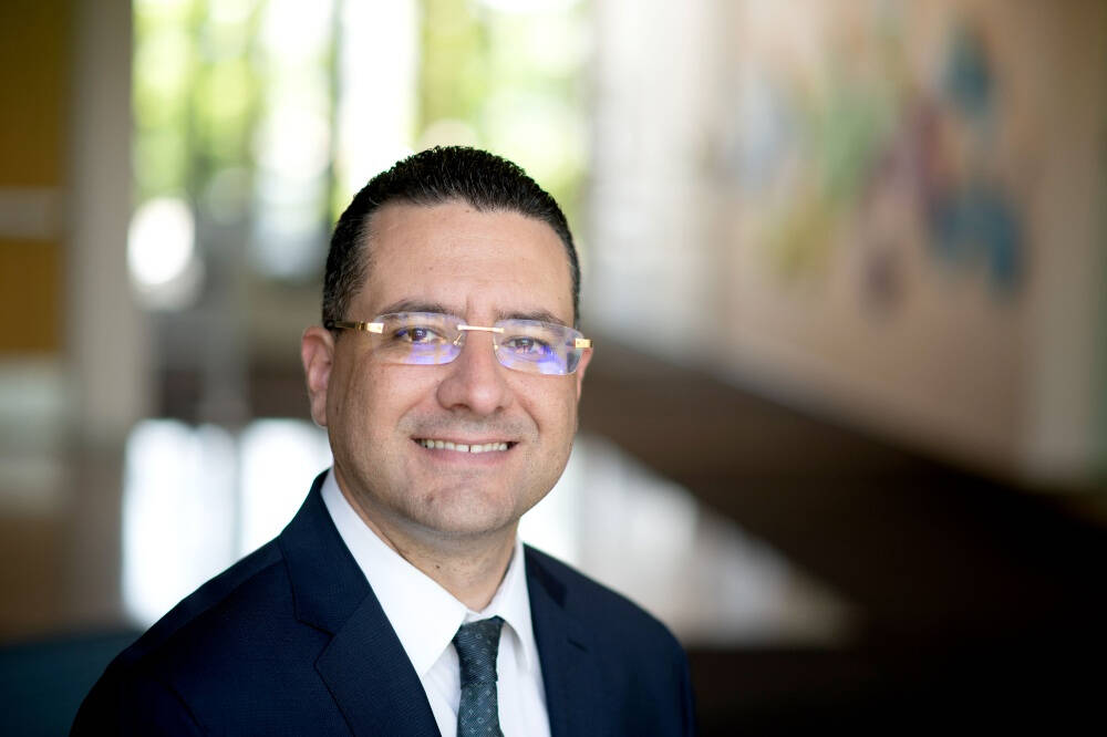 A person with short, dark, neatly styled hair is wearing a dark suit, white shirt, and a dark patterned tie, posed in a modern indoor setting with large windows and abstract wall art in the background.